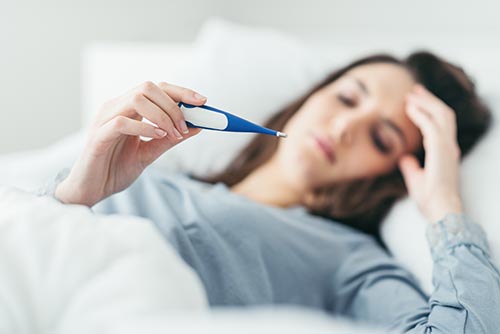 Im Hintergrund unscharf eine brünette Frau im Bett, mit einer Hand hält sie ein Fieberthermometer, das scharf im Vordergrund steht