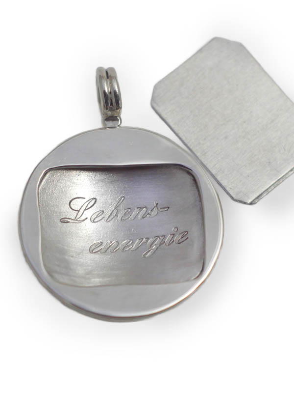 Vor weissem Hintergrund ein rundes, silbernes Amulett, in der Mitte geöffnet, mit dem Wort 'Lebensenergie' eingraviert