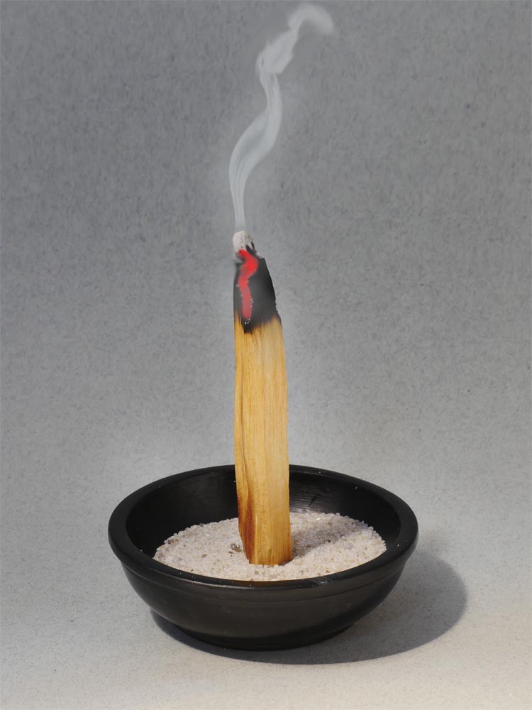 Vor grauem Hintergrung eine schwarze Räucherschale mit Sand, in dem ein glühendes Stück Palo santo Holz steht