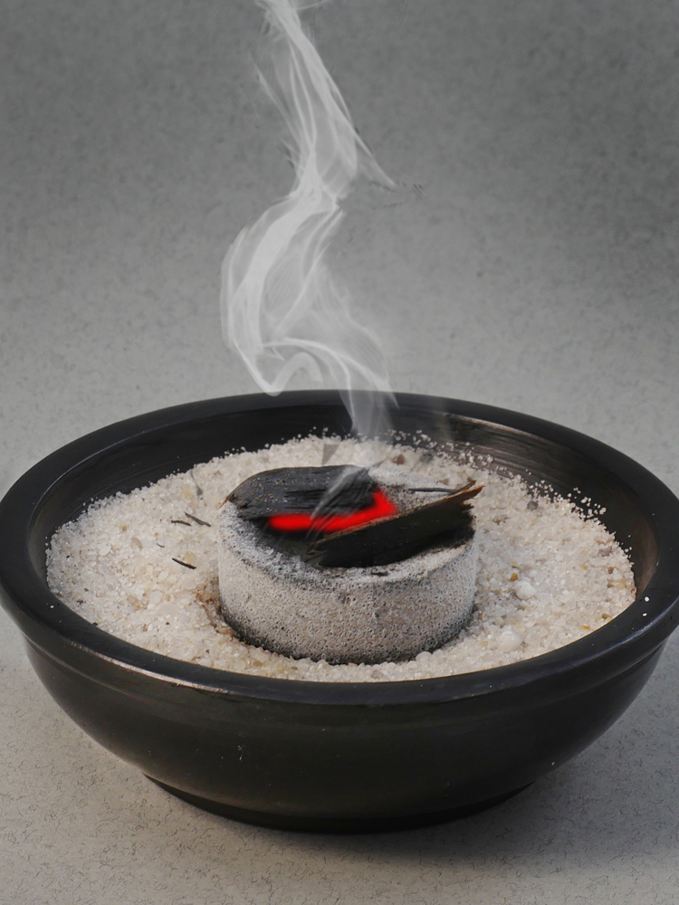 Vor grauem Hintergrund eine schwarze Räucherschale mit grauem Sand, darauf glimmende Kohle, über der Rauch aufsteigt