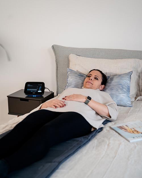 Mit der täglichen Gefäßtherapie BEMER halten Sie sich fit und gesund Eine Frau liegt mit geschlossenen Augen im Bett, unter ihr die graue Ganzkörper-Matte B.Body, neben ihr das BEMER Steuergerät