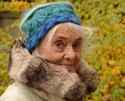 Portrait einer alten Dame mit Pelzkragen und blauer Mütze, im Hintergrund unscharf Herbstlaub