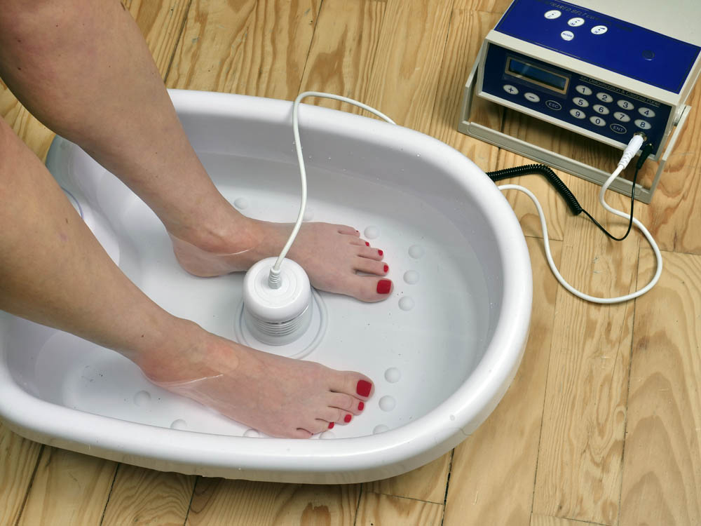 Auf Holzboden steht eine weisse Wanne mit Wasser, darin zwei Füße, daneben ein blauer Kasten