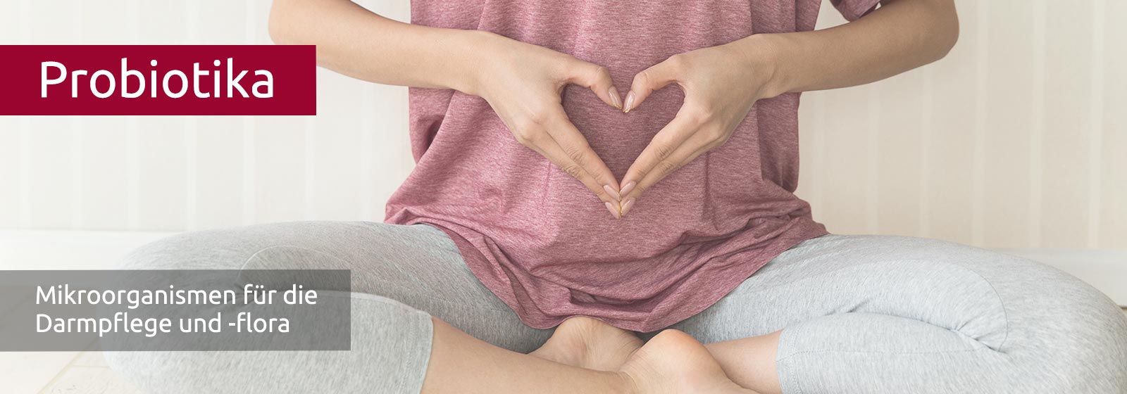 Eine Frau mit grauer Leggins und rosa T-Shirt im Schneidersitz - die Hände formen ein Herz