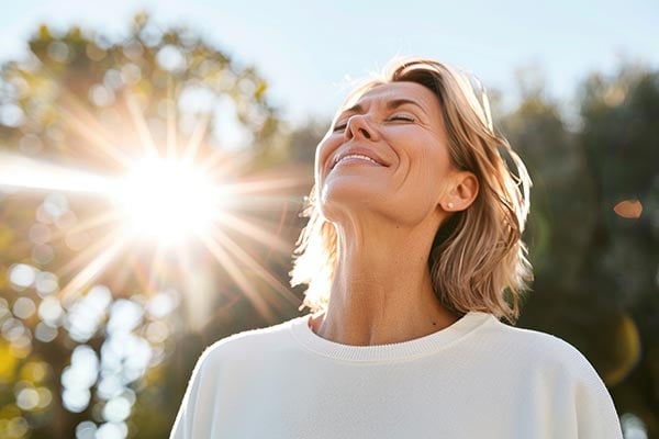 Eine Frau Mitte Vierzig, die lachend den Kopf in den Nacken wirft, links neben ihr Sonnenstrahlen, im Hintergrund unscharf grüne Natur