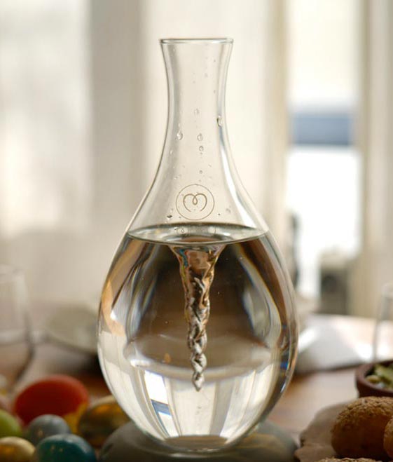 Tropfenförmige Glasflasche mit Wasser gefüllt, auf einem Tisch mit Obst, im Hintergrund unscharf ein Fenster