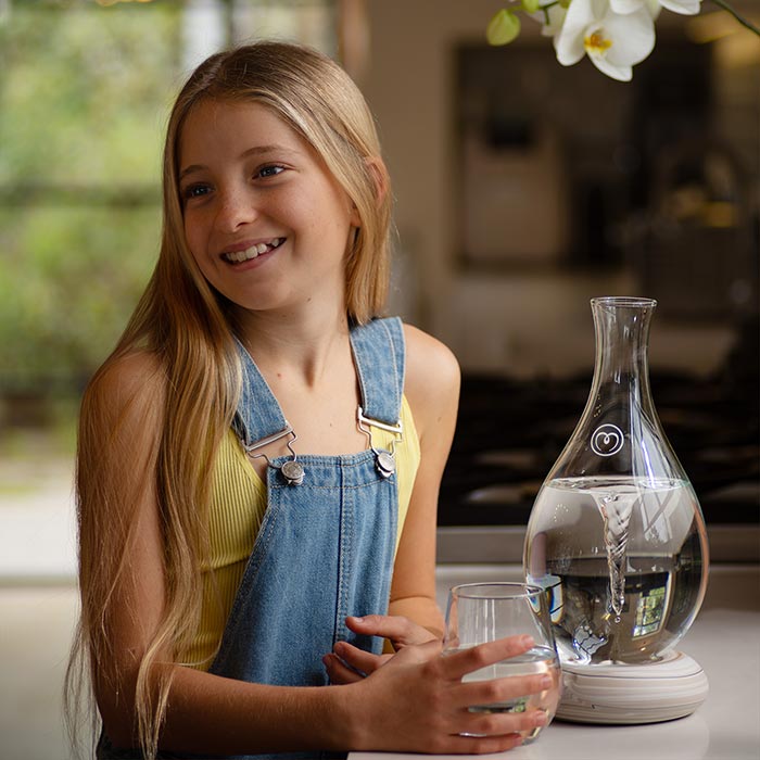 Circa neunjähriges Mädchen mit langen, rotblonden Haaren, das lächelt und ein Trinkglas in der Hand hält, daneben eine schöne, tropfenförmige Glaskaraffe