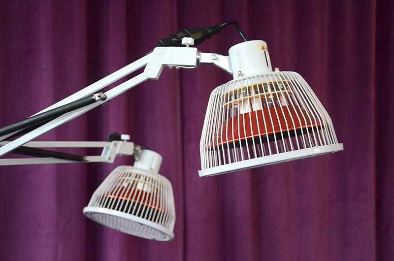 Vorschau: TDP-Lampe Doppelkopf Vorschau: Eine weisse Stehlampe vor lila Hintergrund mit zwei Armen und zwei Therapieköpfen zur Abstrahlung der Infrarot Wärmelampe
